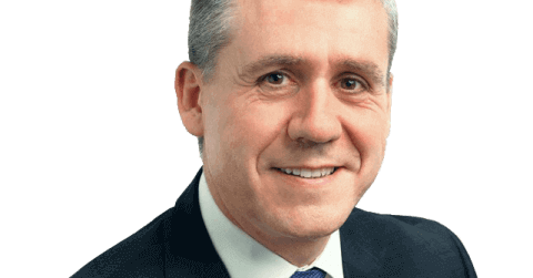 A smiling man in a dark suit with a blue tie and short gray hair stands against a plain white background, embodying the professional elegance of F24 Servicios de Comunicación S.L.U, Spain.