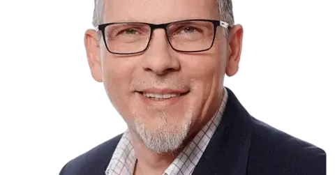 Mann mit kurzen grauen Haaren und Brille, trägt einen dunklen Blazer und ein kariertes Hemd und lächelt vor einem schlichten Hintergrund.
