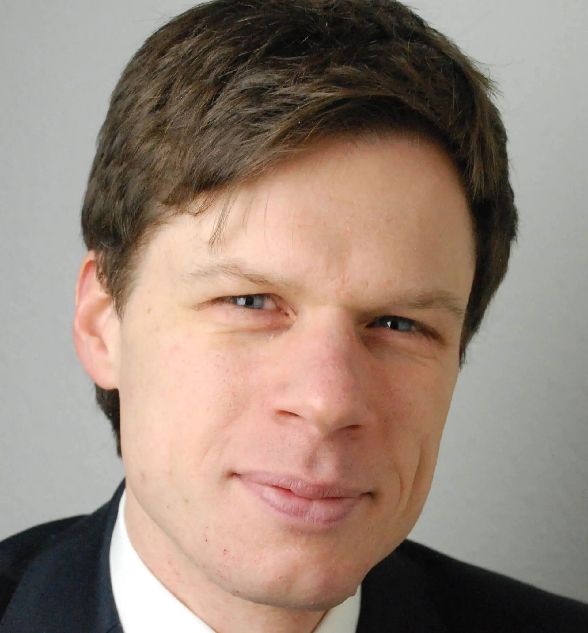 A person with short brown hair is wearing a dark suit and white shirt, looking at the camera with a slight smile, much like a confident speaker poised for a compelling panel discussion at the BCI World Virtual Event 2021. The background is plain gray.