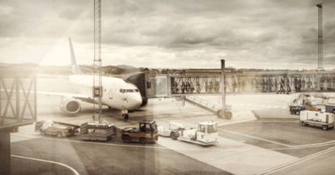 A large airplane is parked at an airport gate, jet bridges connecting to it, with Avinor service vehicles stationed nearby on the tarmac. The overcast sky casts a muted light over the scene, reflective of the question "Hvor høyt er det mulig å nå med CIM?" lingering in the air.