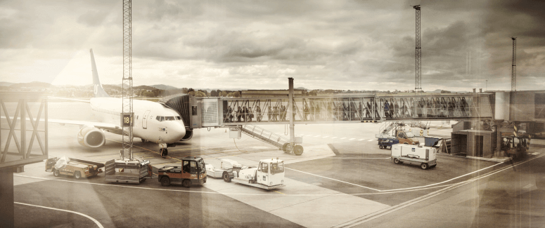 A large airplane is parked at an airport gate, jet bridges connecting to it, with Avinor service vehicles stationed nearby on the tarmac. The overcast sky casts a muted light over the scene, reflective of the question "Hvor høyt er det mulig å nå med CIM?" lingering in the air.