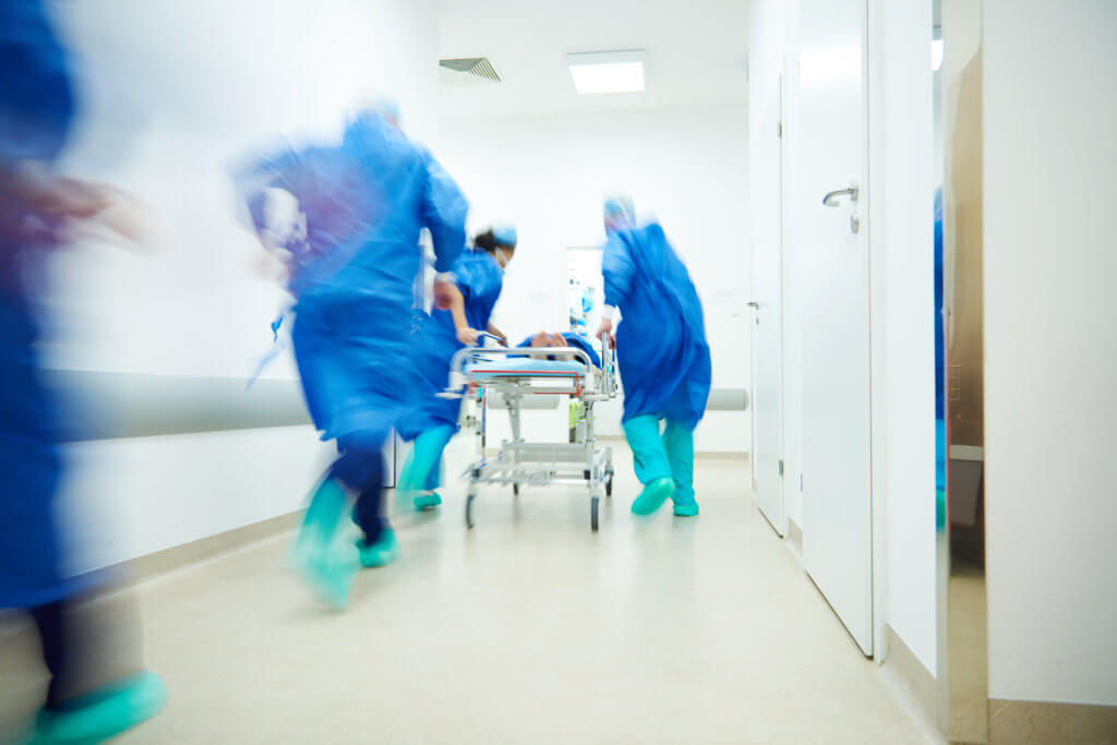 Medical team in blue scrubs and green shoe covers rushes a patient on a stretcher down a hospital corridor. The scene is dynamic, suggesting urgency and critical care. The corridor is brightly lit.