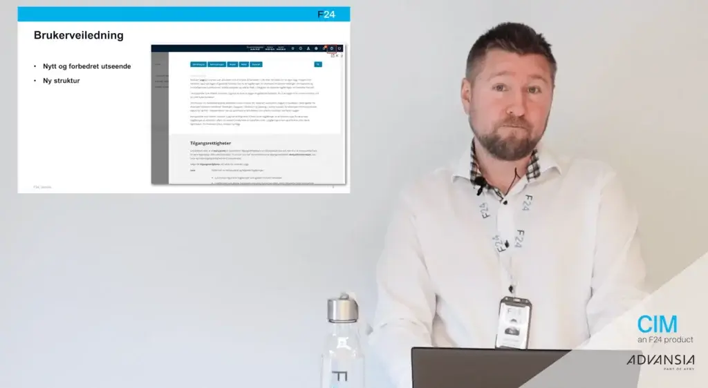 A man stands next to a screen displaying a presentation slide in Norwegian. He's wearing a white shirt and lanyard. The slide includes a webpage screenshot and text about a user manual. A laptop, water bottle, and branding logos are visible.