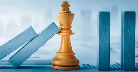 A wooden king chess piece stands upright, stopping a row of blue dominoes from falling, symbolizing Krisenkommunikation amid blurred bar graphs and financial data in the background.