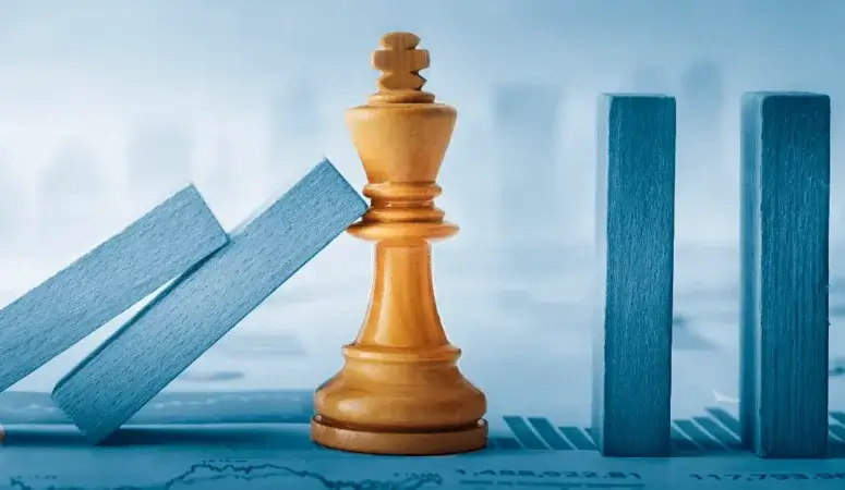 A wooden king chess piece stands upright, stopping a row of blue dominoes from falling, symbolizing Krisenkommunikation amid blurred bar graphs and financial data in the background.