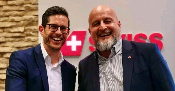 Two smiling men in suits stand in front of a blurred SWISS logo on a wall, embodying the essence of professional Krisenmanagement. One with glasses and dark hair, the other bearded and bald, they exude confidence amidst the light stone accent wall. Icons of FACT24 expertise.
