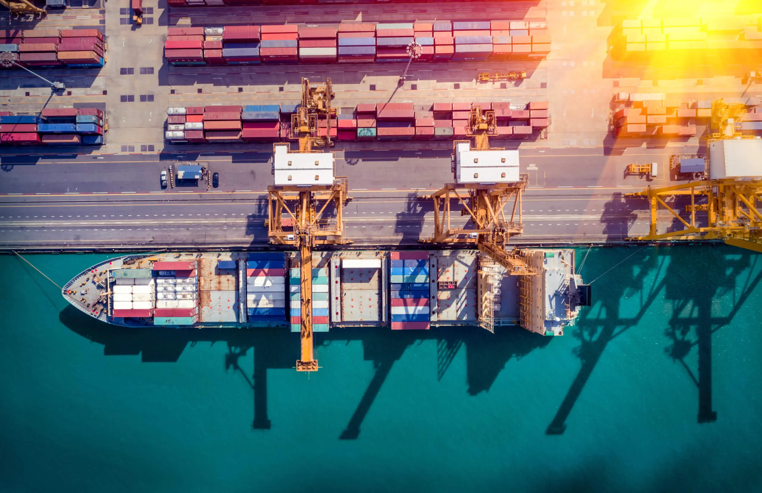 Aerial view of a cargo ship docked at a port. The ship is loaded with colorful shipping containers. Large cranes on the dock are positioned over the ship, and containers are stacked on the ground. The water is a bright turquoise.