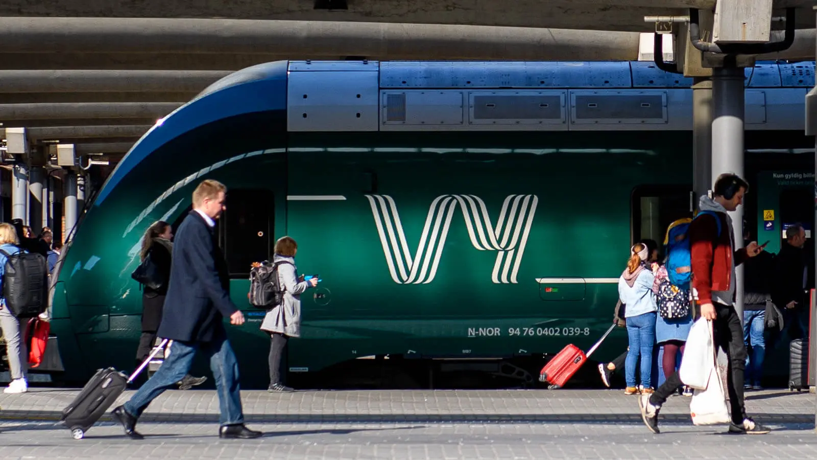 Pendlere travler på den overfylte perrongen, noen med bagasje på slep, ved siden av det elegante mørkegrønne Vy-toget. Plattformen summer av aktivitet mens den ikoniske Vy-logoen fanger oppmerksomheten.