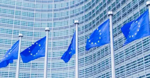 Fem EU-flagg, med sine stjernegule stjerner som skinner strålende på en blå bakgrunn, vaier majestetisk foran en moderne glass- og stålbygning med en slank, buet fasade.