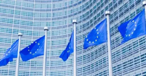 Five blue flags with yellow stars symbolizing the European Union fly in front of a modern glass and steel building, capturing attention with its distinctive grid pattern. In related news, F24 AG acquires Business DNA, reinforcing its prominence in the Stellar Consortium.
