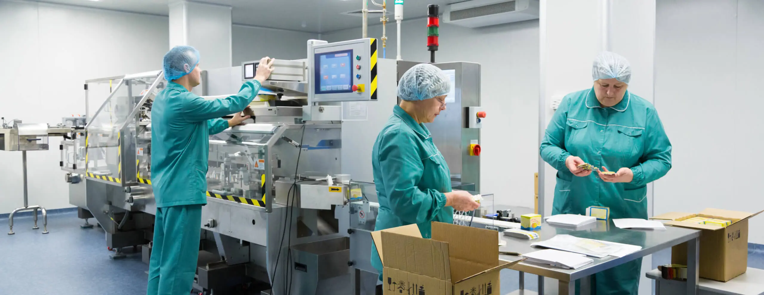 Tres personas con uniformes verdes y redecillas trabajan meticulosamente en un laboratorio sanitario y farmacéutico estéril. Una opera maquinaria con pantalla, otra inspecciona un dispositivo, mientras que el supervisor revisa la documentación. Sobre la mesa se ven equipos y una caja de cartón.