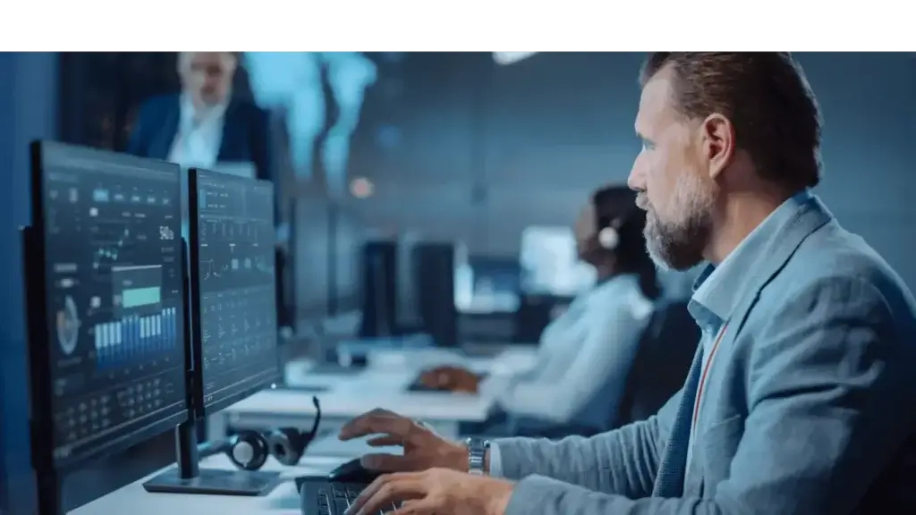 Un homme en costume bleu clair travaille devant plusieurs écrans d'ordinateur affichant des données dans un bureau moderne. Une silhouette floue en arrière-plan et une autre personne portant un casque contribuent à l'animation de l'environnement.