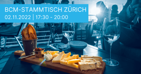 Eine Käseplatte mit verschiedenen Käsesorten, Brot und Aufstrichen steht neben Weingläsern auf dem Tisch beim BCM Stammtisch Zürich. Ein Schild mit der Aufschrift „02.11.2022 | 17:30 - 20:00“ lädt zum Verweilen ein. Im Hintergrund sitzen Menschen, die vom Sonnenlicht durchflutet werden.