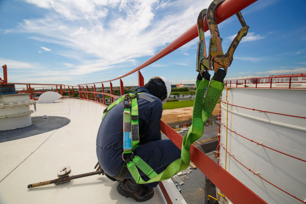 Accroupi sur une passerelle industrielle, un ouvrier vêtu d'un équipement de protection incarne l'engagement envers la protection du travail, maintenu par un harnais de sécurité fixé à une rampe rouge. Le décor, un grand réservoir de stockage et le ciel à perte de vue, soulignent l'importance de la sécurité, en particulier pendant l'« Alleinarbeit ».