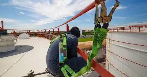 A worker in safety gear kneels on an elevated platform, demonstrating Occupational Health and Safety practices with green harnesses secured to a red railing. The sky is blue with clouds, and industrial tanks are visible in the background.