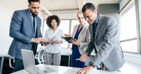 På et lyst, moderne kontor er fire fagfolk forankret i å diskutere hvordan teste beredskapsplanen. Tre står mens en sitter ved bordet. Kledd i forretningsantrekk bruker de bærbare datamaskiner og nettbrett blant spredte papirer, badet i naturlig lys som strømmer gjennom store vinduer.
