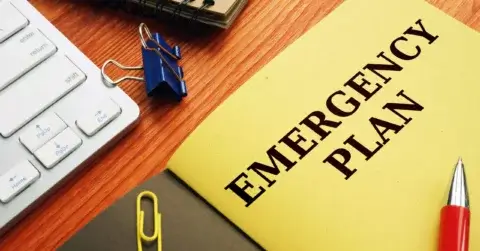 En gul mappe merket "Emergency Plan" sitter på et trepult ved et tastatur, en lukket notatbok med en blå permklips og en rød penn. Like ved holder beredskapstiltak-mappen sølv og gul binders.