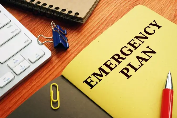 En gul mappe merket "Emergency Plan" sitter på et trepult ved et tastatur, en lukket notatbok med en blå permklips og en rød penn. Like ved holder beredskapstiltak-mappen sølv og gul binders.