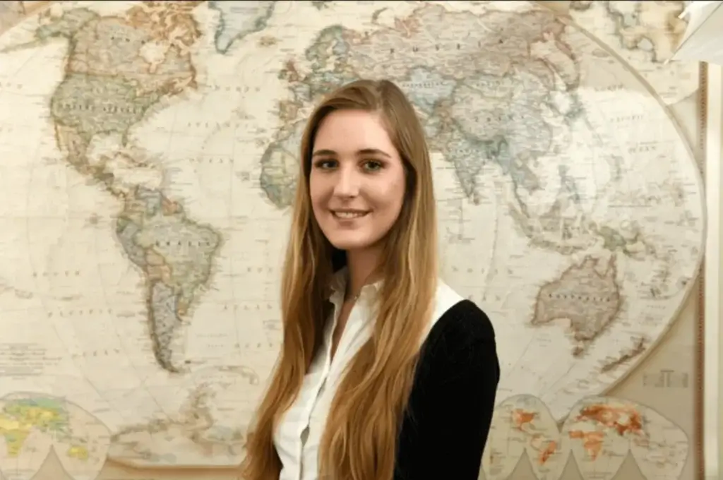 A person with long hair stands smiling in front of a large, detailed world map, their excitement as vibrant as the colors and labels that span continents and countries. They are wearing an ROS-logo black top over a white shirt, perfectly complementing the global theme.