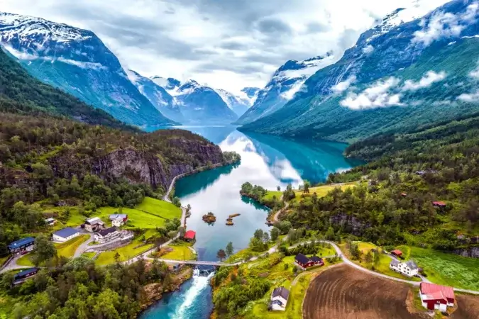 En naturskjønn luftfoto av en rolig fjord omgitt av frodige grøntområder og snødekte fjell, som minner om det fredelige landskapet som ble sett under innreiseregistrering. Sjarmerende landlige hus og jorder ligger ved det rolige turkise vannet, med skyer som driver rundt toppene.