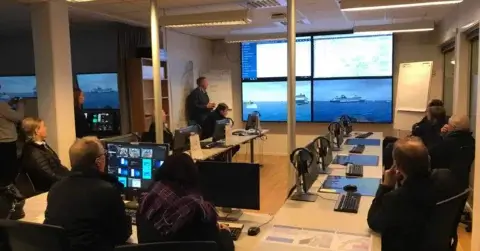Et kontrollrom med personer som sitter ved skrivebord utstyrt med datamaskiner. De observerer en presentasjon på flere store skjermer som viser maritime scener og data. Rommet er godt opplyst, og det er aktiv diskusjon blant deltakerne.