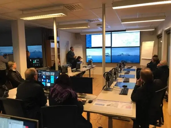 Et kontrollrom med personer som sitter ved skrivebord utstyrt med datamaskiner. De observerer en presentasjon på flere store skjermer som viser maritime scener og data. Rommet er godt opplyst, og det er aktiv diskusjon blant deltakerne.