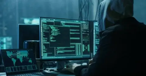 En person som har på seg en hettegenser sitter foran flere dataskjermer og viser kode, digitale kart og ROS-simuleringer. Den svakt opplyste innstillingen antyder et hemmelighetsfullt eller hackende miljø, med en stasjonær datamaskin og en bærbar datamaskin som viser global kartgrafikk.
