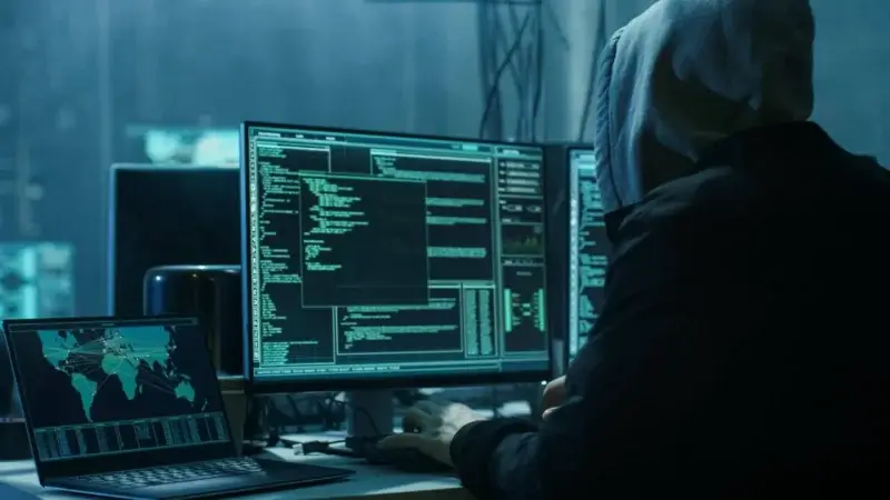 En person som har på seg en hettegenser sitter foran flere dataskjermer og viser kode, digitale kart og ROS-simuleringer. Den svakt opplyste innstillingen antyder et hemmelighetsfullt eller hackende miljø, med en stasjonær datamaskin og en bærbar datamaskin som viser global kartgrafikk.