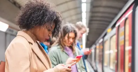 Folk står på en T-baneplattform, engasjerte med smarttelefonene sine, kanskje leser en varsling mens de venter på toget til høyre. Det buede taket over legger til den urbane scenen hvor individer er absorbert i enhetene sine og sømløs hendelseshåndtering utfolder seg rundt dem.