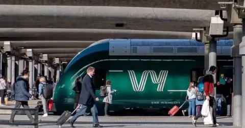 Et grønt tog utsmykket med den karakteristiske "Vy"-logoen står ved en travel stasjonsplattform. Folk vever seg gjennom mengden, bærende poser, mens andre dveler i nærheten av toget. Overhead spor kaster intrikate mønstre av skygger på den travle plattformen nedenfor.