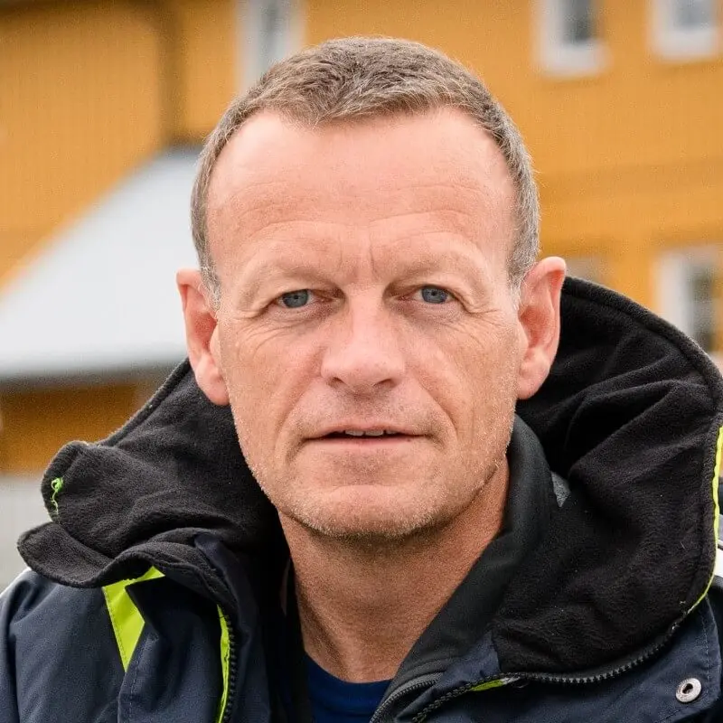 A person with short hair wearing a jacket with a hood and yellow accents stands confidently in front of a yellow building. The background is slightly blurred, highlighting their thoughtful expression as if formulating a crisis plan.