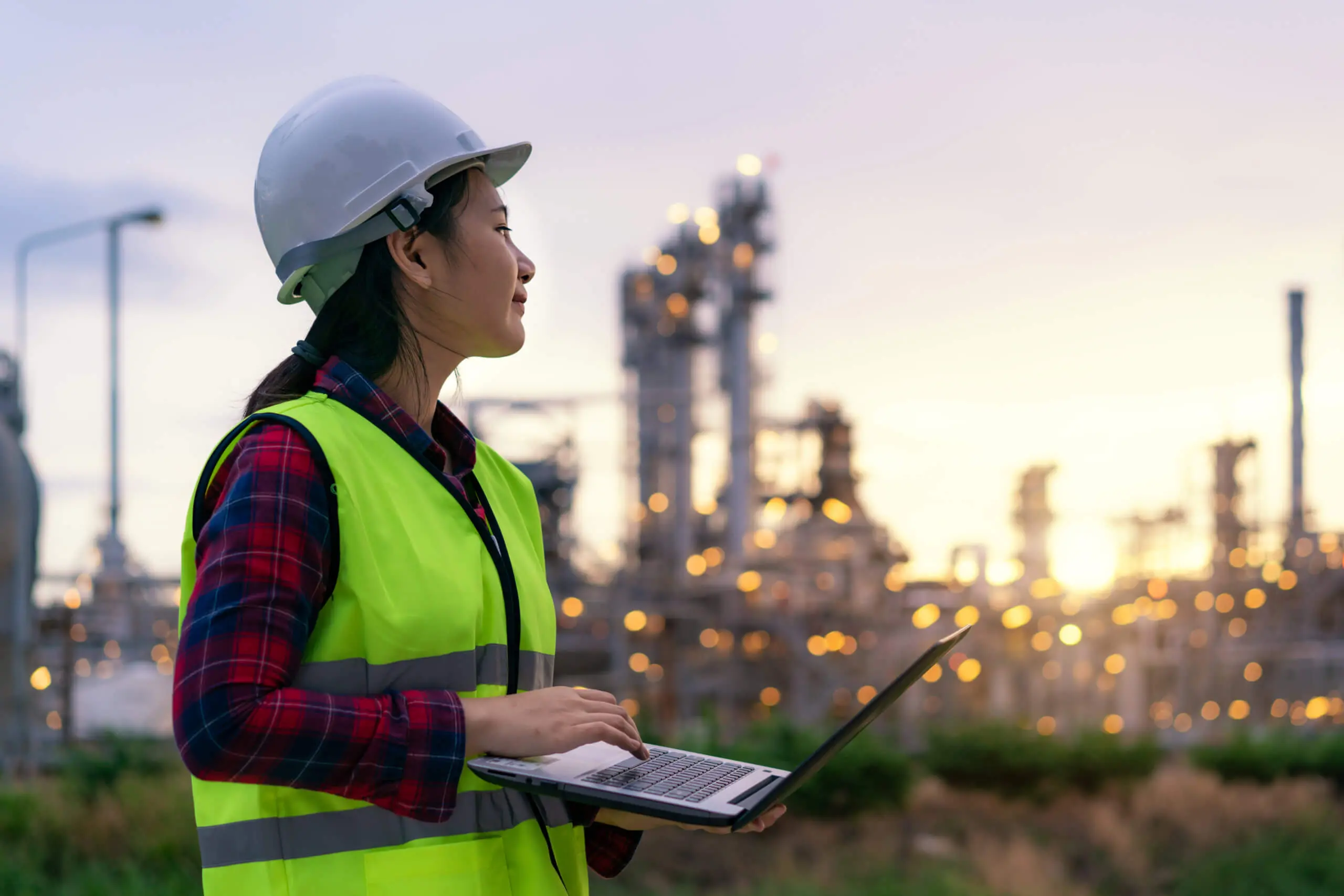 Eine Person mit Schutzhelm und Warnweste steht mit einem Laptop im Freien und überwacht die intelligenten Sicherheitssysteme der Industrieanlage. Im Hintergrund funkeln die verschwommenen Lichter der Anlage und sorgen für die reibungslose technische Alarmierung in der Dämmerung.