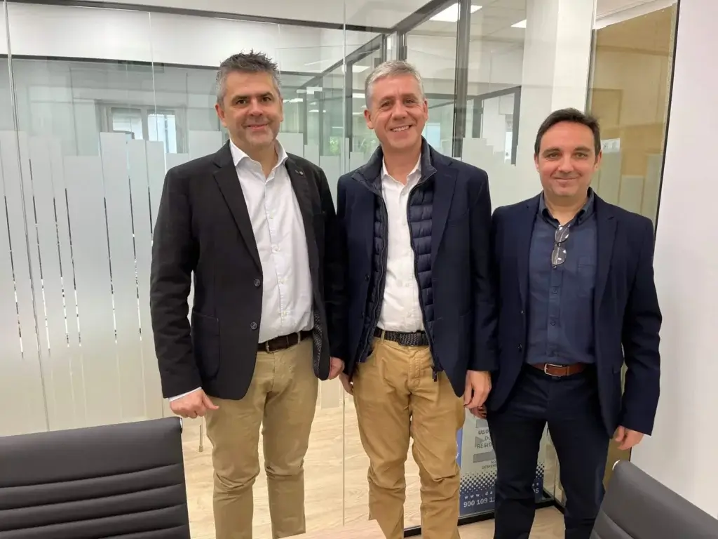 Three men in business attire stand and smile in an office with glass walls, symbolizing how F24 strengthens its market position in Spain. Positioned by a conference table, the room exudes professionalism with its neutral color scheme and abundant natural light.