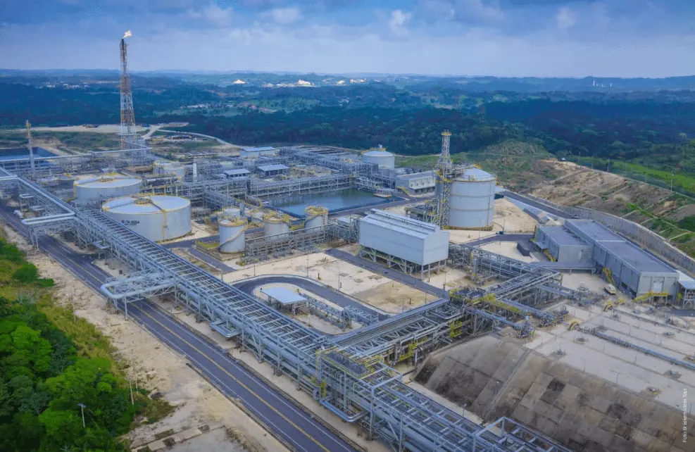 Vista aérea de un gran complejo industrial con múltiples estructuras metálicas interconectadas, tanques de almacenamiento y tuberías dedicadas a la preparación de desastres naturales, todo situado en un paisaje montañoso y verde bajo un cielo azul con nubes.