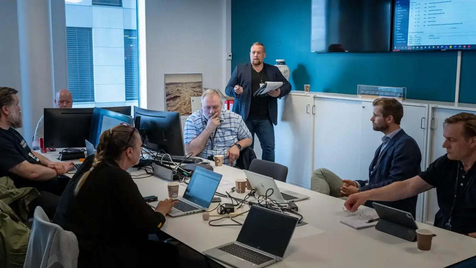 En gruppe mennesker er samlet i et møterom for en workshop med Audun Martinsen, deres bærbare datamaskiner åpne rundt bordet. Én person står foran og holder en notisblokk, mens en skjerm viser informasjon på veggen med blågrønn aksent, i kontrast til de elegante hvite møblene.