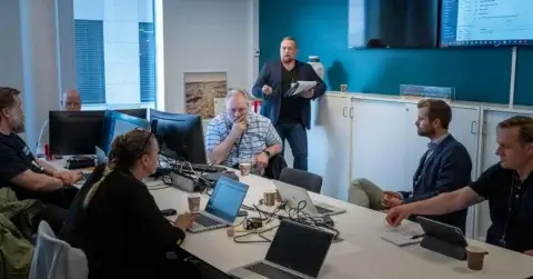 En gruppe mennesker er samlet i et møterom for en workshop med Audun Martinsen, deres bærbare datamaskiner åpne rundt bordet. Én person står foran og holder en notisblokk, mens en skjerm viser informasjon på veggen med blågrønn aksent, i kontrast til de elegante hvite møblene.