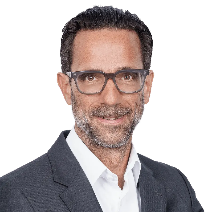 A man with dark hair and a trimmed beard is wearing glasses, a white shirt, and a dark suit jacket. He is smiling, looking slightly to the side, embodying the professional spirit of F24 Schweiz AG in Switzerland. The background is plain.