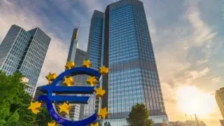 Un grand symbole bleu de l'euro orné d'étoiles jaunes se dresse fièrement devant un groupe de grands immeubles de bureaux modernes au coucher du soleil, soulignant l'importance de la gestion des risques pour les institutions financières. Le ciel partiellement nuageux et le soleil chaud illuminent cette scène de F24.com, avec des arbres visibles à gauche.