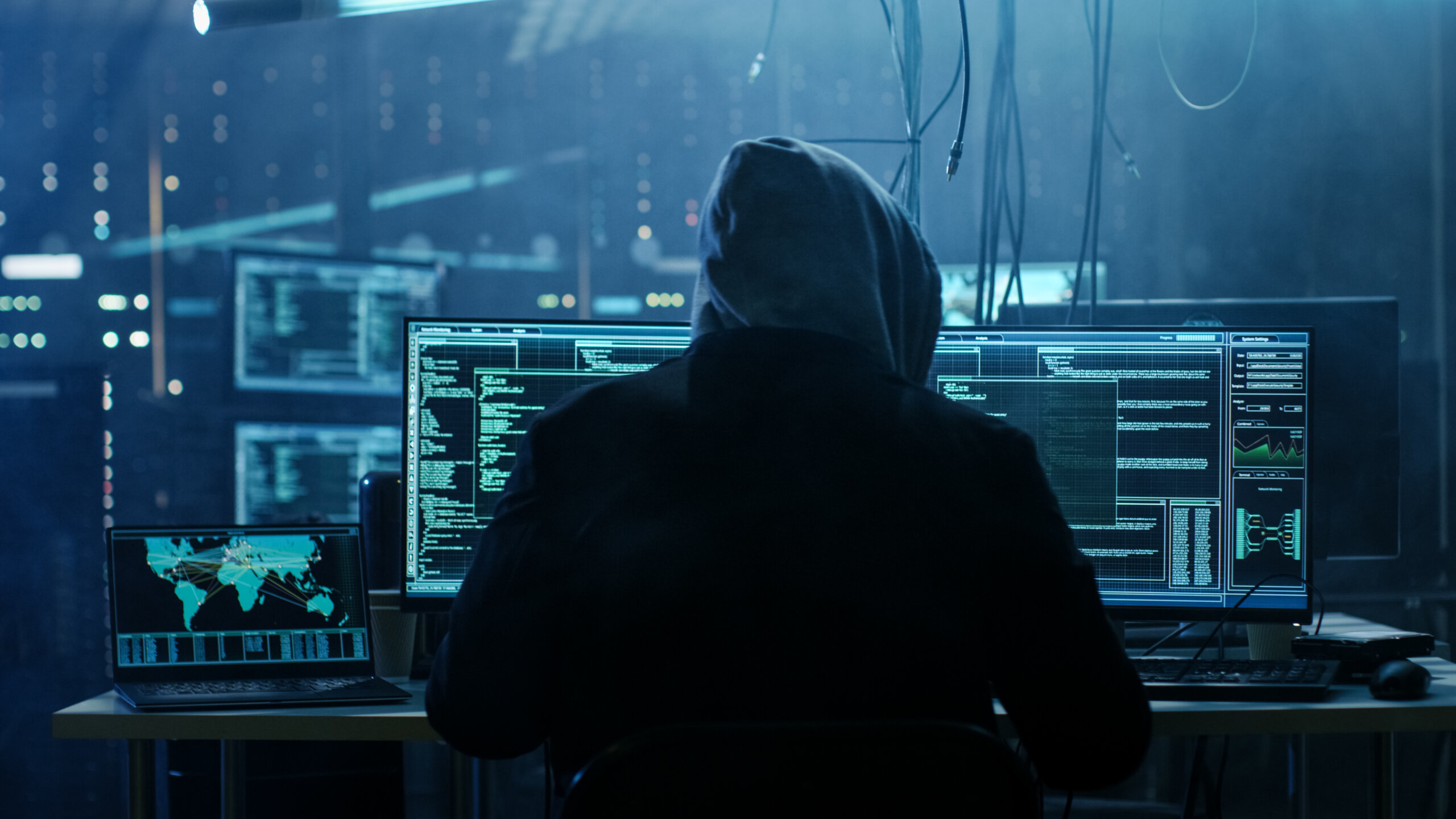 Una persona con una sudadera con capucha está sentada ante un escritorio con varios monitores en los que se muestran códigos y datos, en una habitación a oscuras, lo que sugiere un entorno de ciberseguridad o piratería informática centrado en la respuesta ante incidentes cibernéticos.