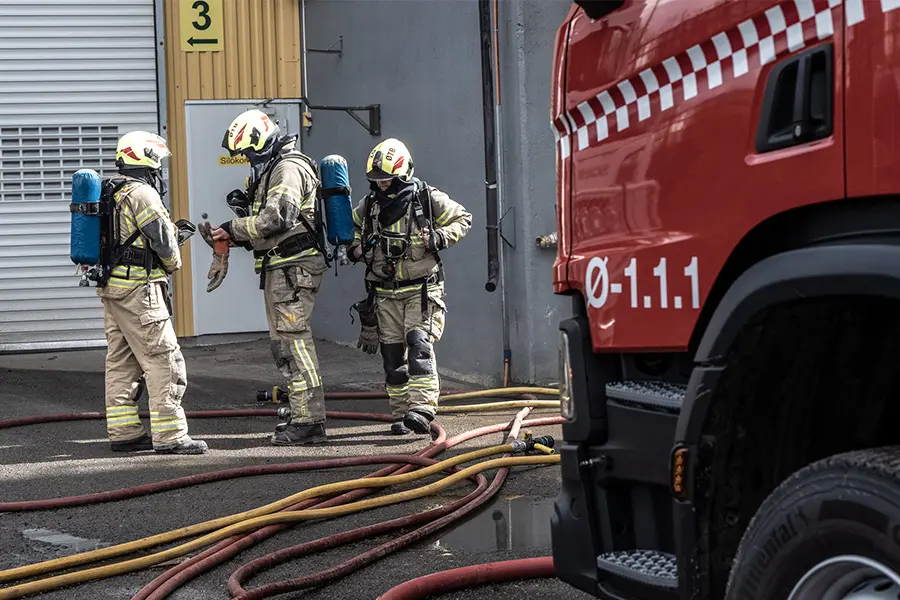 Tre brannmenn i fullt utstyr og hjelmer står ved brannslangene utenfor en bygning, med en rød brannbil i forgrunnen. CIM sikrer kvalitet og kontroll innen brann og redning mens de diskuterer eller justerer utstyret.