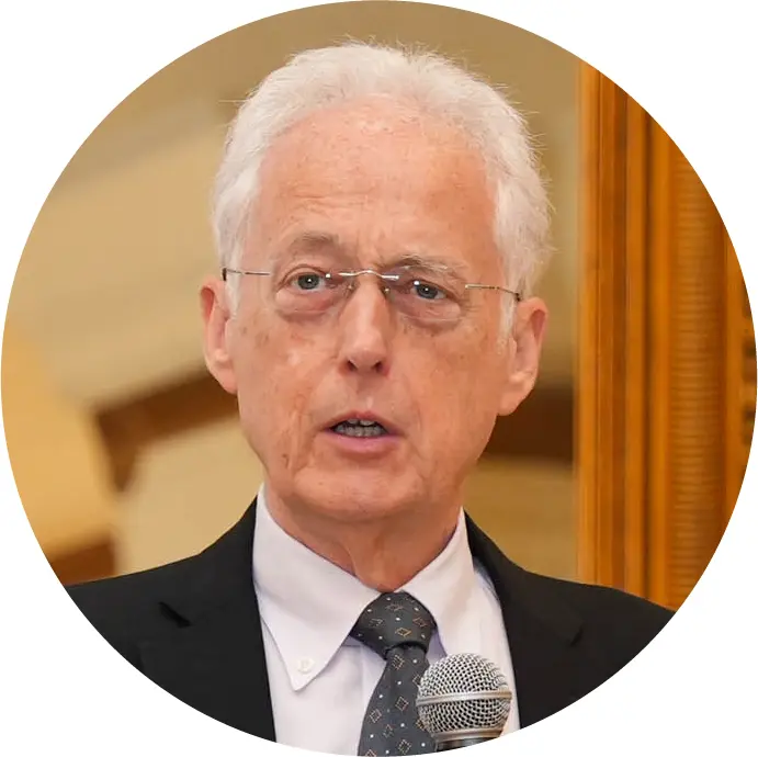 An older man with white hair and glasses, wearing a suit and tie, speaks into a microphone at an event. The softly blurred background suggests an indoor setting focused on organisational resilience.