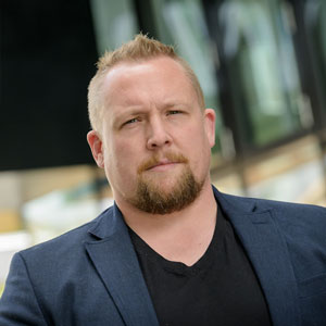 A man with short blond hair and a beard, wearing a navy blazer over a black shirt, stands outdoors with a serious expression. The background is blurred with modern glass buildings.