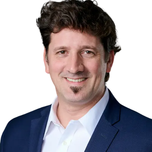 A man with wavy brown hair and facial hair is smiling, wearing a white shirt and a dark blue blazer. The background is plain white.