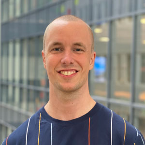 A smiling person with a shaved head wearing a navy blue shirt with thin, multicolored stripes stands indoors in front of a large windowed building.