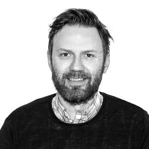 A man with a beard and mustache smiles at the camera. He has short, slightly messy hair and is wearing a plaid shirt under a dark sweater. The background is plain white.