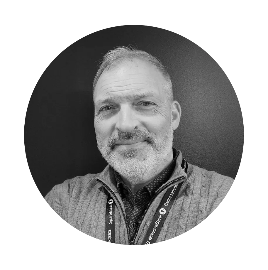 Black and white close-up of a smiling man with short hair and a beard, wearing a jacket and lanyard. The background is plain and dark.