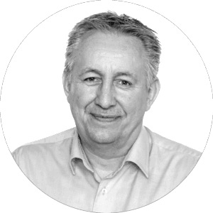 A grayscale portrait of an older man with short, light hair, smiling and wearing a light-colored collared shirt, centered in a circular frame.