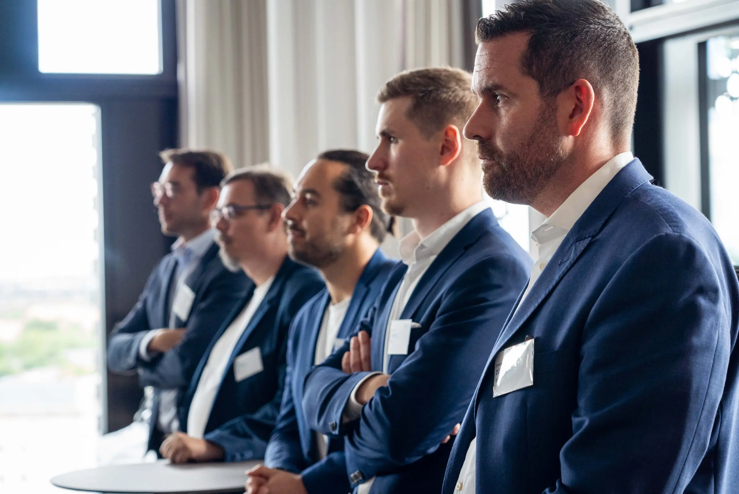 A group of five men in business attire stand in a line, all facing the same direction. They are indoors, with natural light streaming from large windows behind them. Each wears a suit with a name tag on their jacket, attending the F24 Experience Tour München – an event for resilience and crisis management.