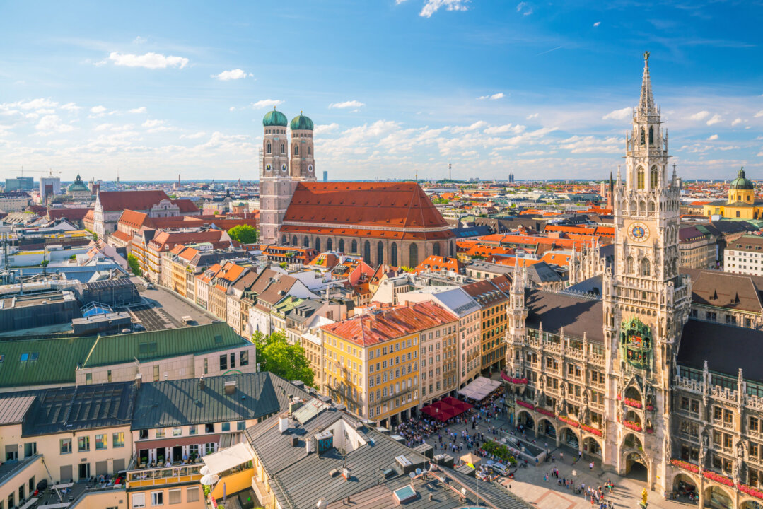 Aerial view of central Munich, Germany, featuring the red-roofed Frauenkirche with twin towers and the ornate New Town Hall (Neues Rathaus) on Marienplatz, surrounded by colorful historic buildings under a blue sky.