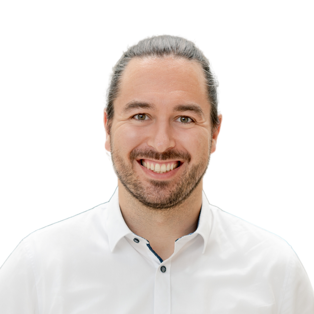 Un homme souriant aux longs cheveux bruns tirés en arrière en chignon et à la barbe taillée, portant une chemise blanche boutonnée, pose sur un fond blanc uni.